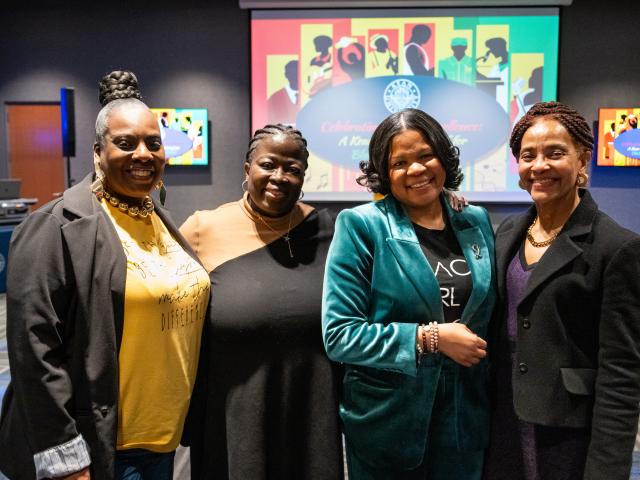 A group of four people standing together and posing for a photo in an event space. They are dressed in semi-formal attire, and a colorful presentation slide with silhouettes and bold patterns is displayed on the screen behind them. The room has patterned carpeting and dim lighting, with additional screens and event materials visible in the background.
