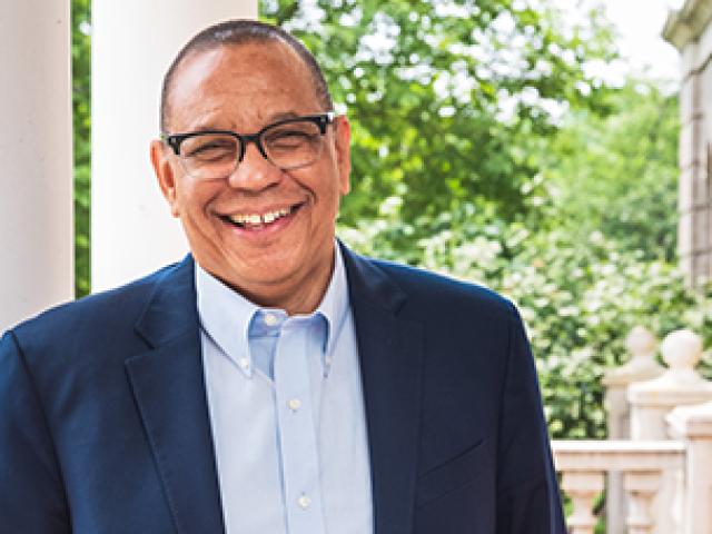 Dr. Dale Caldwell standing and smiling wearing a navy blue suit jacket, blue shirt and black glasses. 