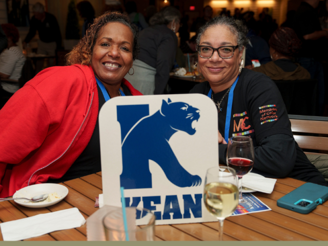 Two women in dimly lit room sitting at table with glasses, plates and Kean signage
